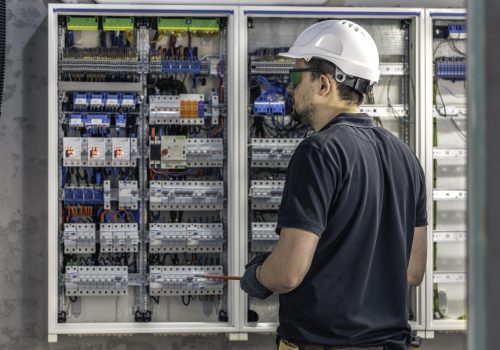 A male electrician works in a switchboard using an electrical connection cable, connecting equipment with tools. View from the back.