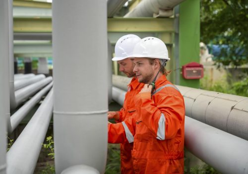 Two male workers wearing hard hats and orange uniforms are examining large pipes at an industrial facility. They appear focused on their task, surrounded by a green environment.